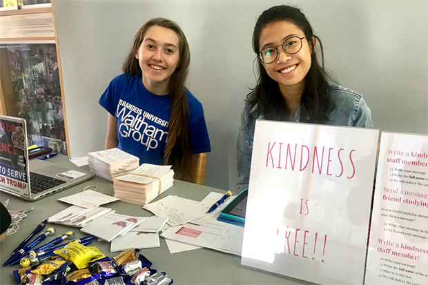 Two students, one wearing a Waltham Group t-shirt smile from behind a table of give aways including cards, candy and pens. A sign reads "Kindness is Free."